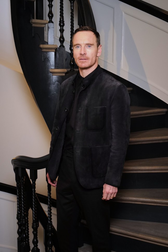 Michael Fassbender wears a black suit as he poses for a photo with a spiral staircase behind him