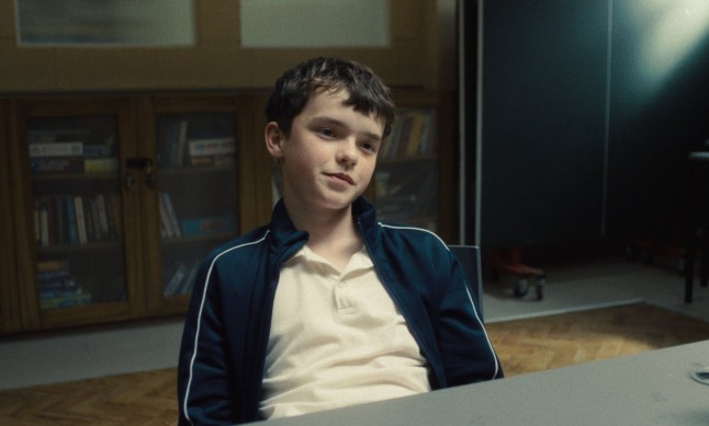Owen Cooper as Jamie Miller in Adolescence. He's sitting at a table with a smug expression on his face.