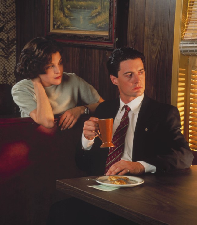 Dake Ciioer sits in a diner booth drinking a cup of coffee while a woman looks over his shoulder.