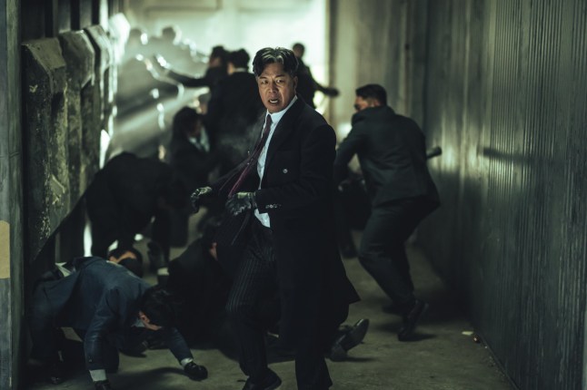 A still from Mercy For None of a man in a suit with men fighting behind him